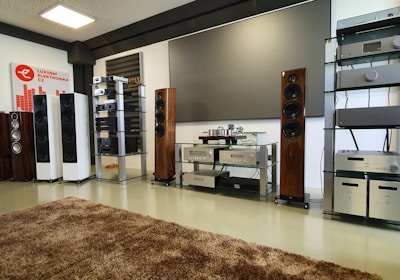 A spacious audio equipment showroom featuring a variety of high-end sound systems. Several tall speakers are aligned against the walls, displaying sleek wooden and metallic finishes. Electronics such as amplifiers and receivers are arranged on modern glass shelves, and a large, plush brown rug covers the floor. A sign on the left side suggests a focus on luxury electronics.