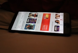 A smartphone displaying a streaming app interface, showing various movie and TV show thumbnails. The screen includes categories like 'For you' and 'New release hit films', with images from popular films. The background is a dimly lit setting with a fabric surface.
