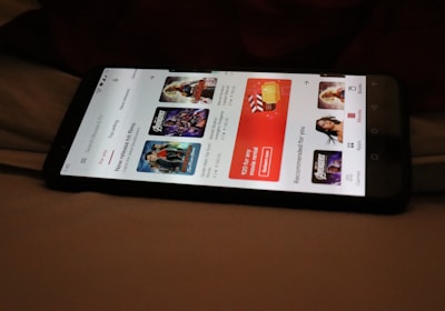 A smartphone displaying a streaming app interface, showing various movie and TV show thumbnails. The screen includes categories like 'For you' and 'New release hit films', with images from popular films. The background is a dimly lit setting with a fabric surface.