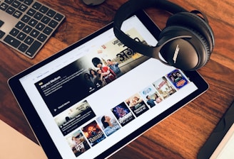 A wooden table holds a tablet displaying a streaming service interface with various movie and TV show thumbnails. Nearby, a pair of Bose headphones rests on the table along with a keyboard and a mouse.