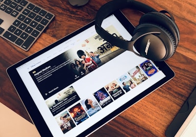 A wooden table holds a tablet displaying a streaming service interface with various movie and TV show thumbnails. Nearby, a pair of Bose headphones rests on the table along with a keyboard and a mouse.