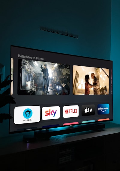 A television screen displaying a user interface with popular streaming service apps including Netflix, Sky, Apple TV, Prime Video, and others. The screen also shows images from movies or shows above the apps. The room is dimly lit with a potted plant visible on the side.