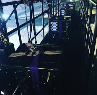 A narrow, industrial-looking platform with a series of large, black equipment cases secured with straps. Several control panels with glowing blue lights are visible on the front of each case. The platform is enclosed with metal railings, and various cables and chains hang along the sides, adding to the technical and utilitarian atmosphere. Bright lighting from behind casts a dramatic contrast against the darker sections of the platform.