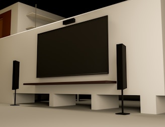 A modern home entertainment setup featuring a large flat-screen TV mounted on a light-colored wall. Two tall black speakers stand on either side of the TV. The room has a minimalist design with a clean, uncluttered look. Shelving and a wooden door are visible in the background.
