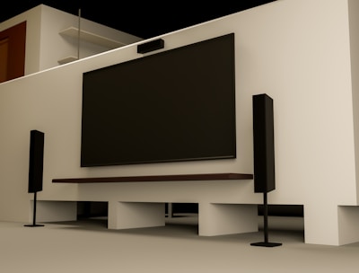 A modern home entertainment setup featuring a large flat-screen TV mounted on a light-colored wall. Two tall black speakers stand on either side of the TV. The room has a minimalist design with a clean, uncluttered look. Shelving and a wooden door are visible in the background.
