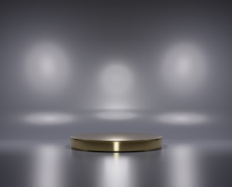 A metallic platform is centered in a minimalistic setting with a reflective surface. Soft, diffused lighting creates circular patterns on the background wall, enhancing the focus on the central stage. The ambiance is sleek and modern, with a hint of elegance.